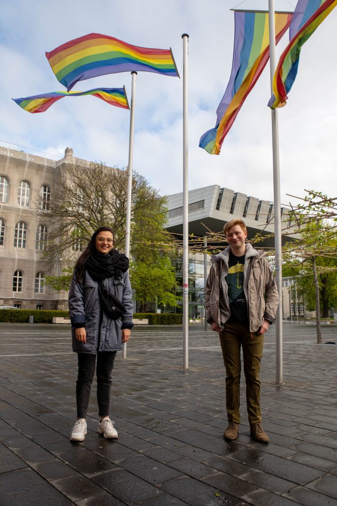 Die beiden Beauftragten vor dem Hauptgebäude mit Regenbogenflaggen die im Hintergrund wehen. Aline steht links und Jan rechts.